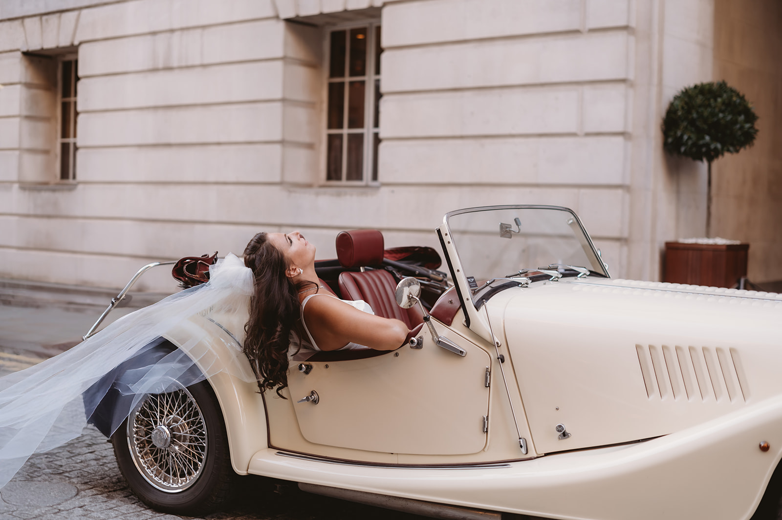 Bride in an ivory Morgan 4/4 with her wedding veil flowing behind the car